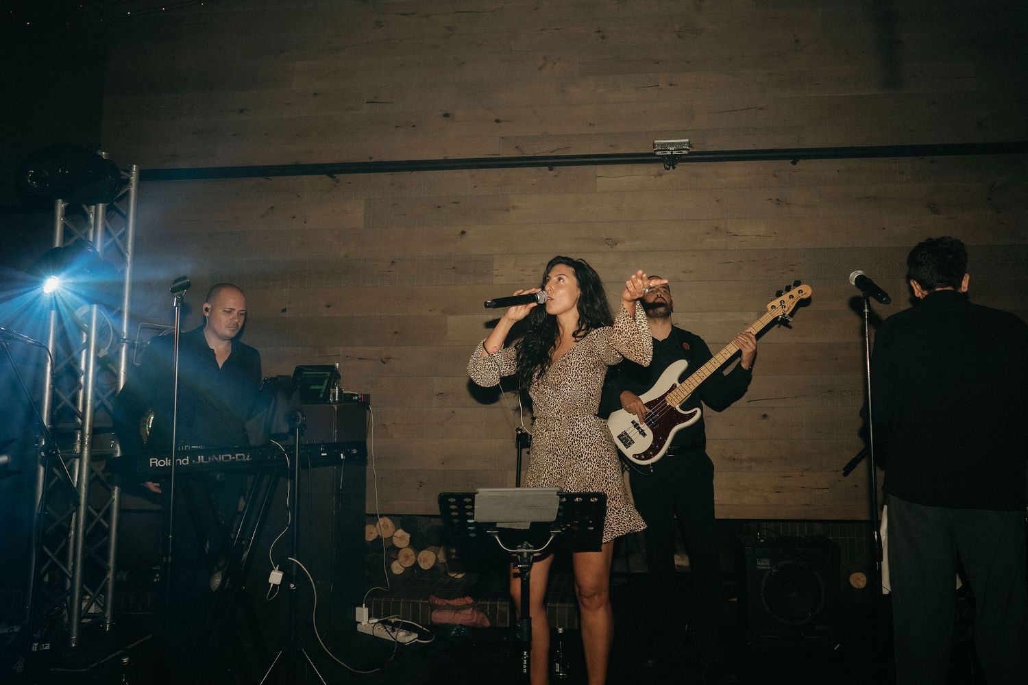 A band performs on stage with musicians playing instruments against a wooden wall backdrop in moody lighting.