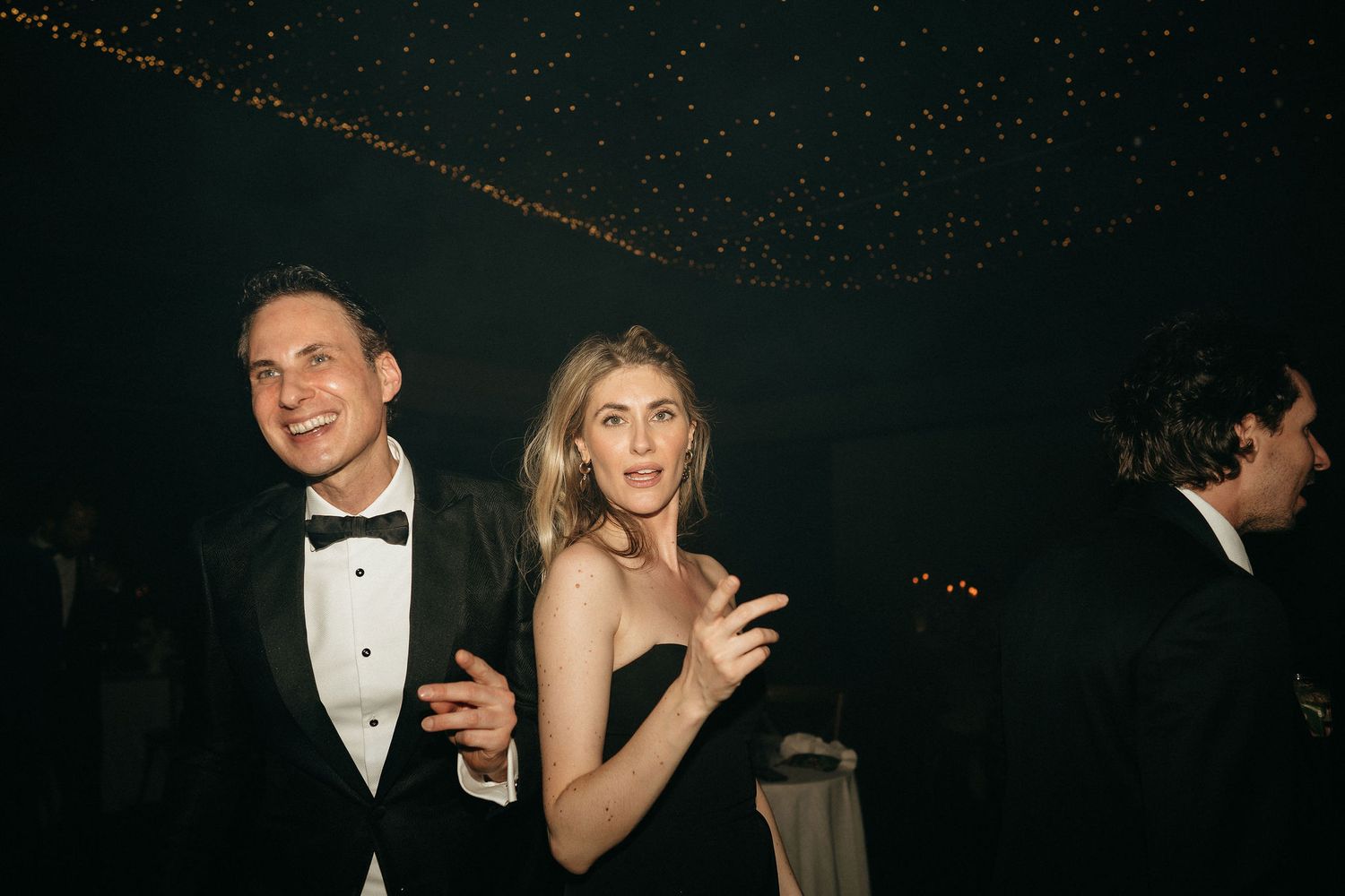 Partygoers in formal attire enjoying themselves at an evening celebration with twinkling lights overhead.
