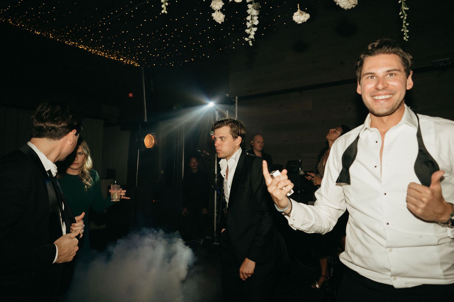 People dancing and celebrating at a dimly lit wedding reception with hanging lights creating a festive atmosphere.
