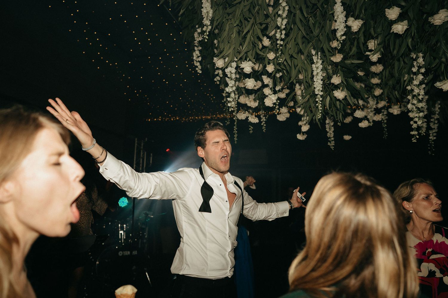 Wedding guests dancing together on a dark dance floor illuminated by overhead party lights.