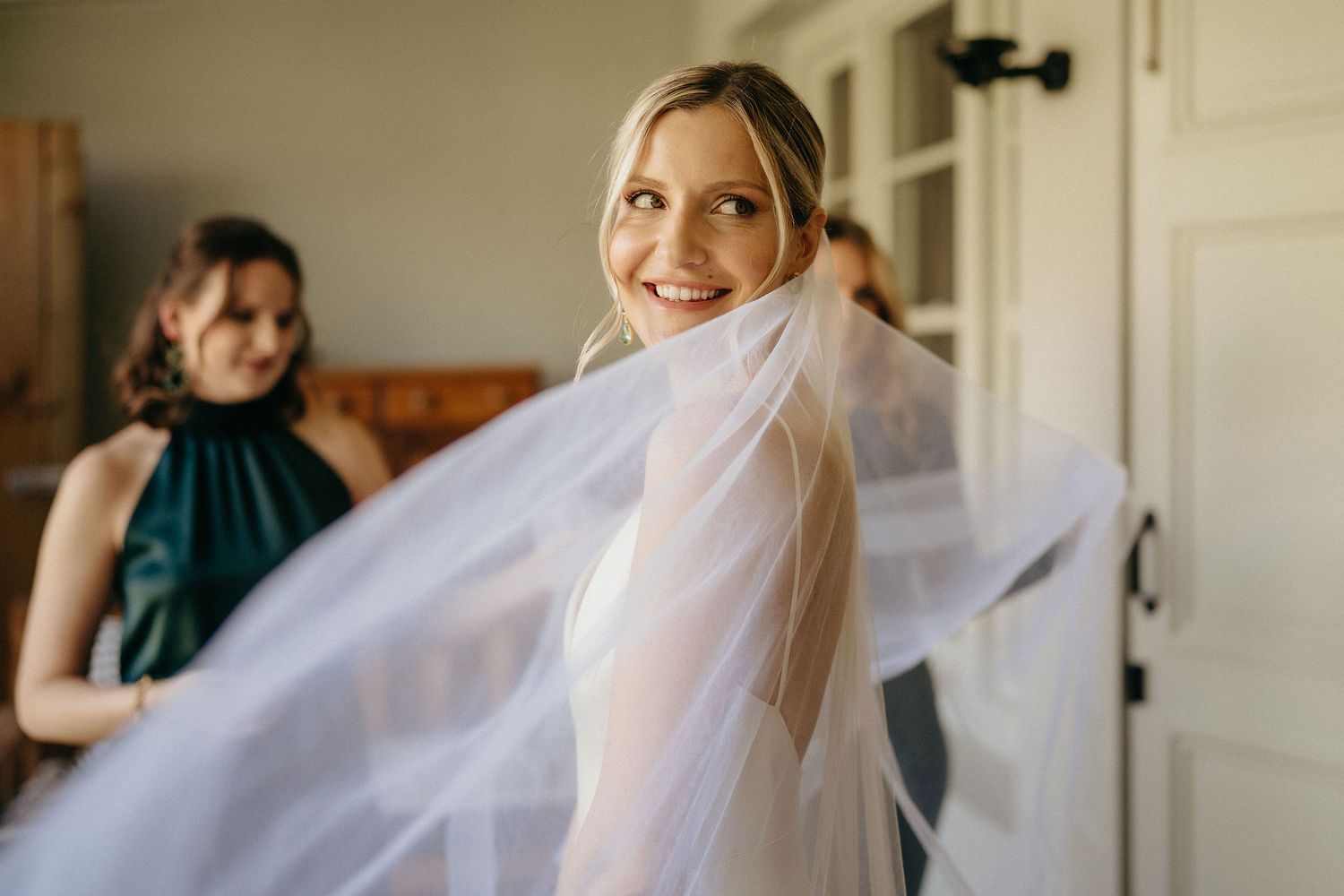 A bride in a white wedding dress and veil stands in front of a white door during wedding preparations.