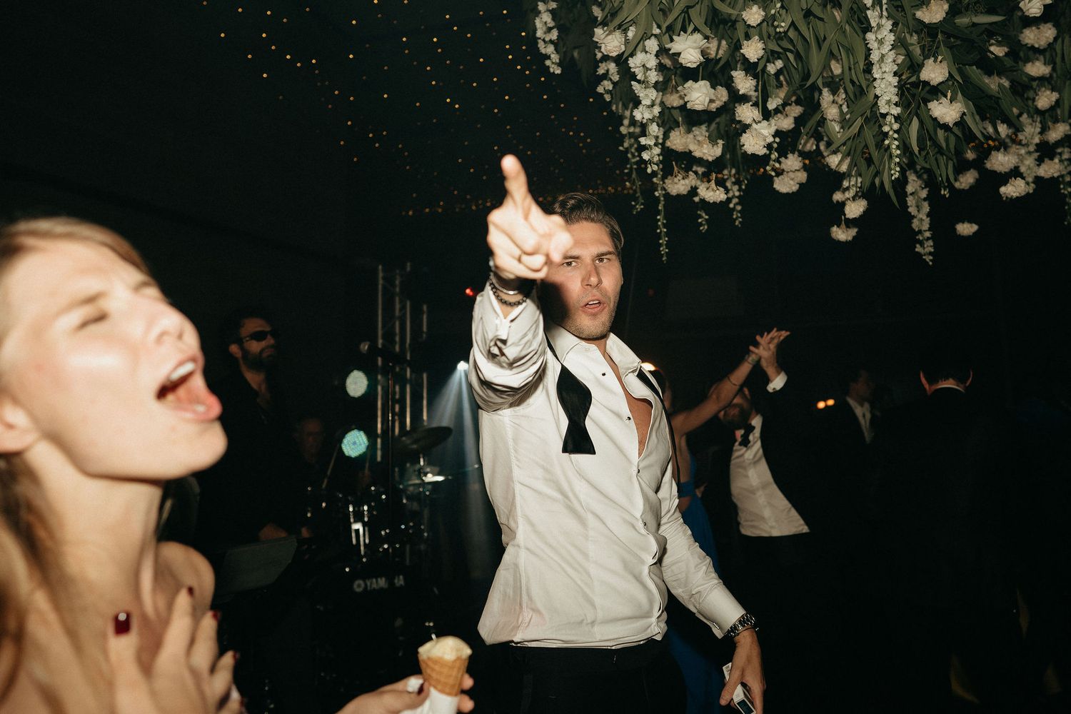 People dancing and celebrating at a dark nightclub with someone in a white shirt pointing upward on the dance floor.