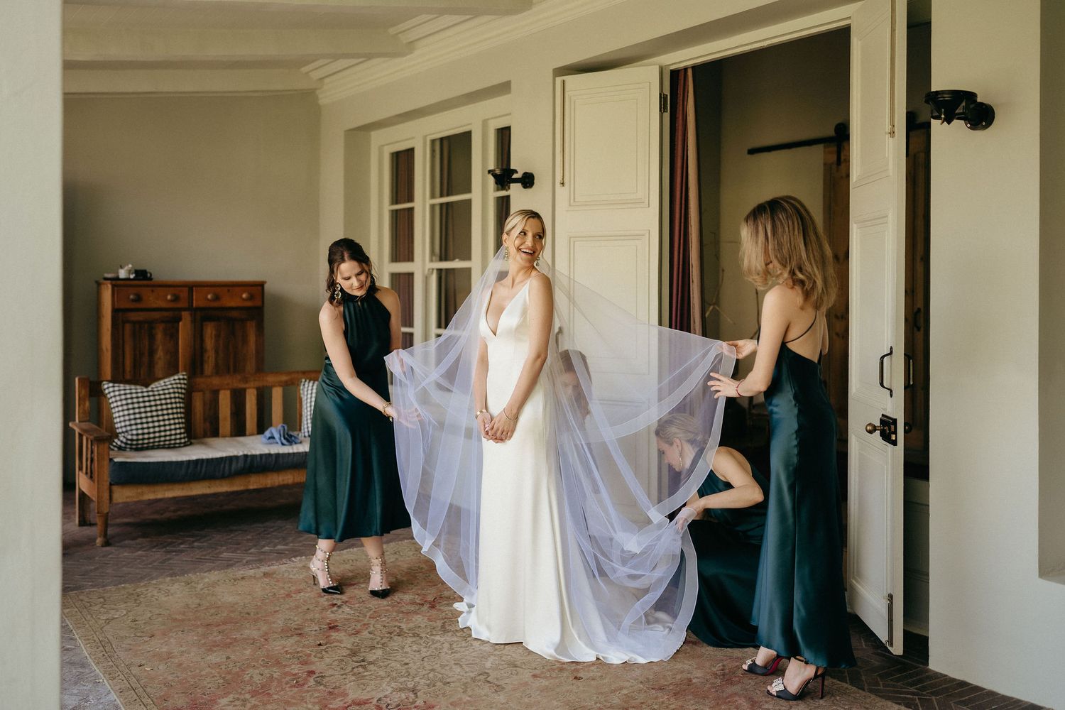 A panoramic view of a bride and bridesmaids in teal dresses preparing in a bright hallway.