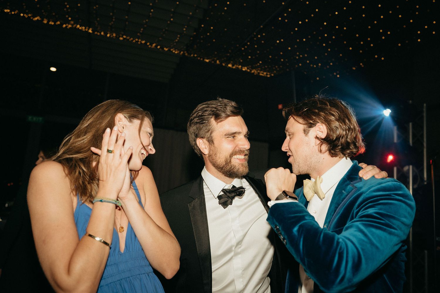 Three people share a joyful moment together on a dance floor with string lights twinkling in the background.