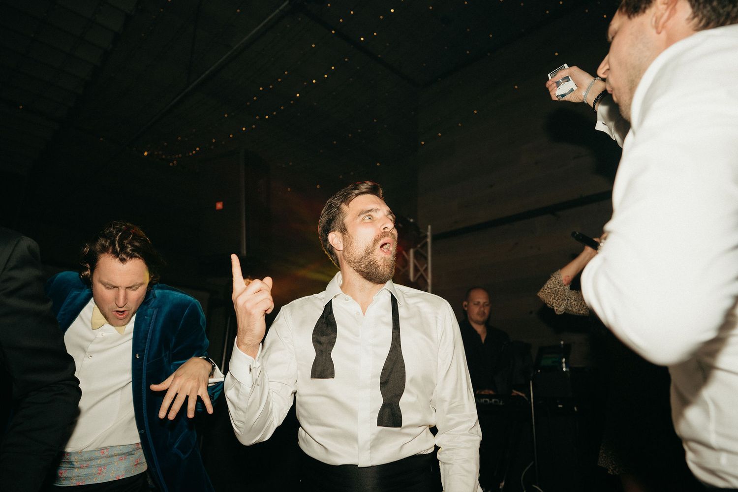 People dancing energetically at a nighttime party with one person gesturing upward in a white dress shirt and tie.