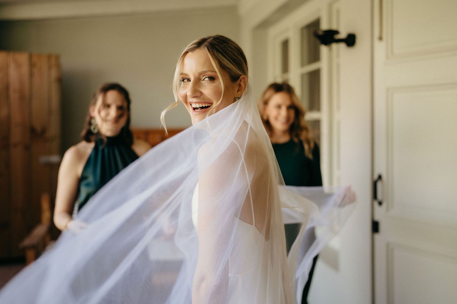 A bride in a flowing white gown and veil gets ready on her wedding day in a sunlit room.