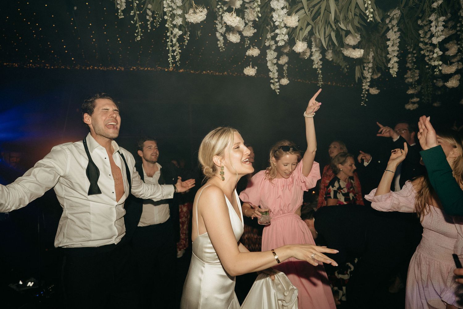 People dancing together at a dimly lit party or wedding reception with hanging decorative lights.