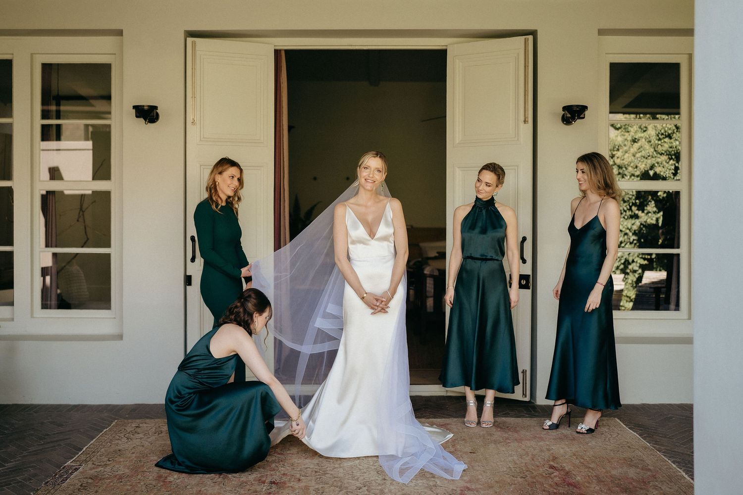 A row of bridesmaids in dark teal dresses stand with a bride in white on a covered porch.