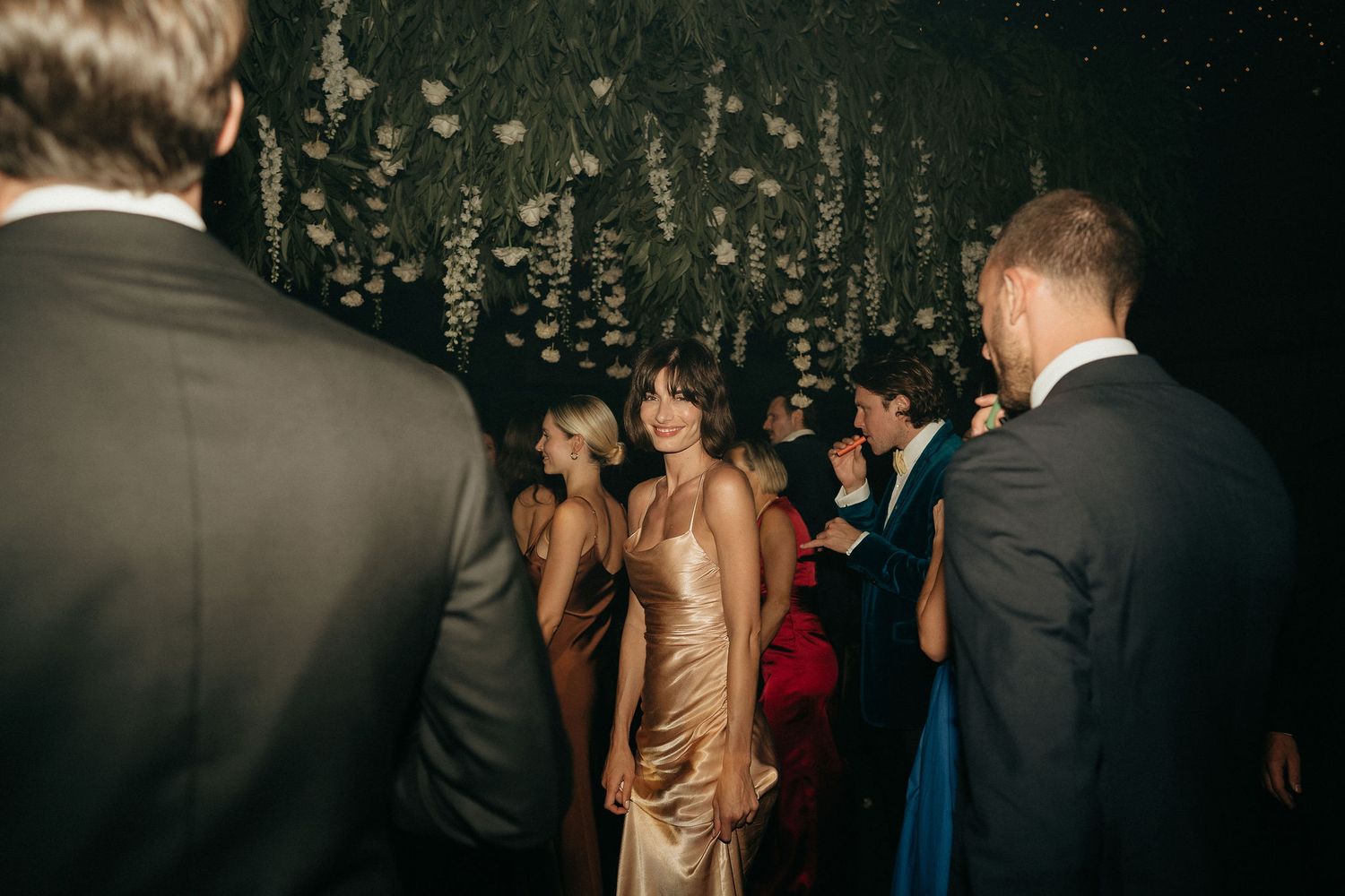 Wedding guests dancing and socializing on a dimly lit dance floor beneath elegant hanging floral decorations.