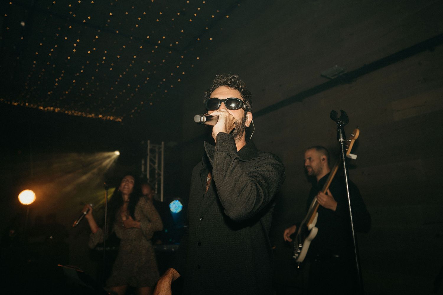 A performer in sunglasses sings into a microphone on a dark stage with dramatic lighting and band members in the background.