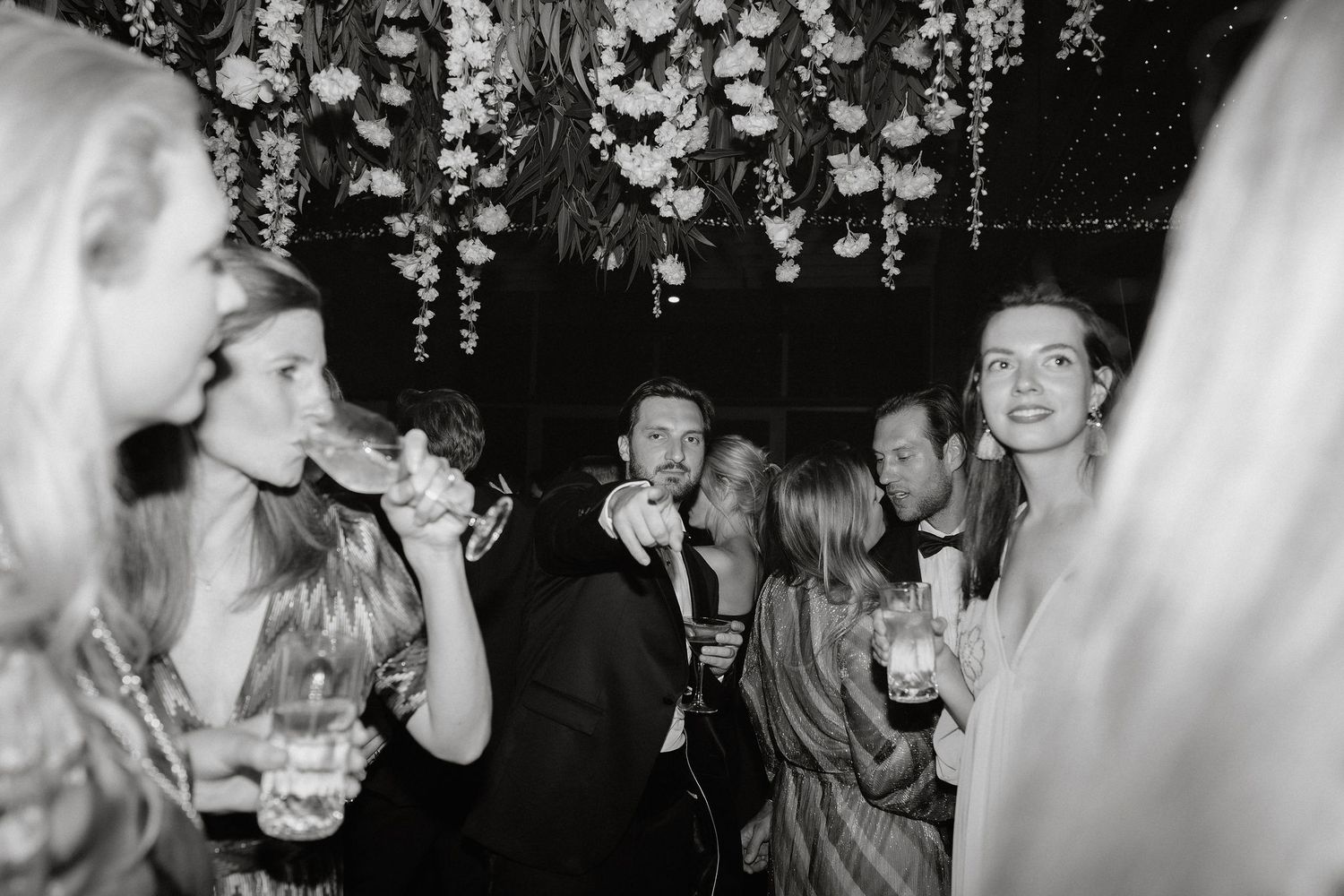 Wedding reception guests dance and celebrate on a dimly lit dance floor under hanging floral decorations.