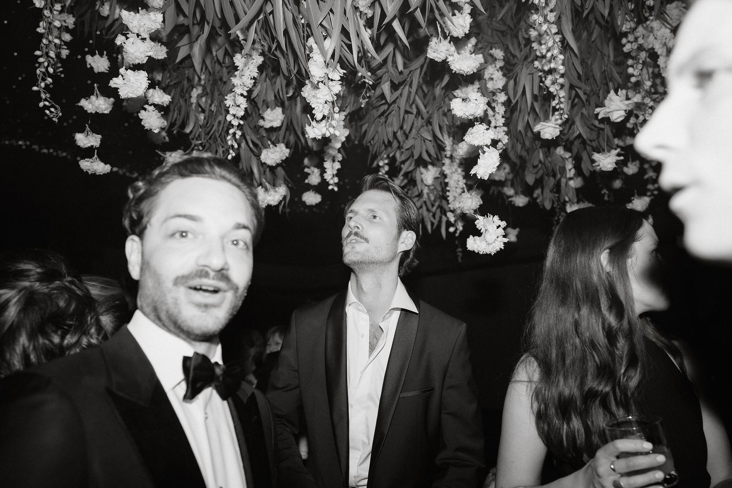 A black and white party photo showing guests in formal attire under hanging floral decorations at an elegant event.