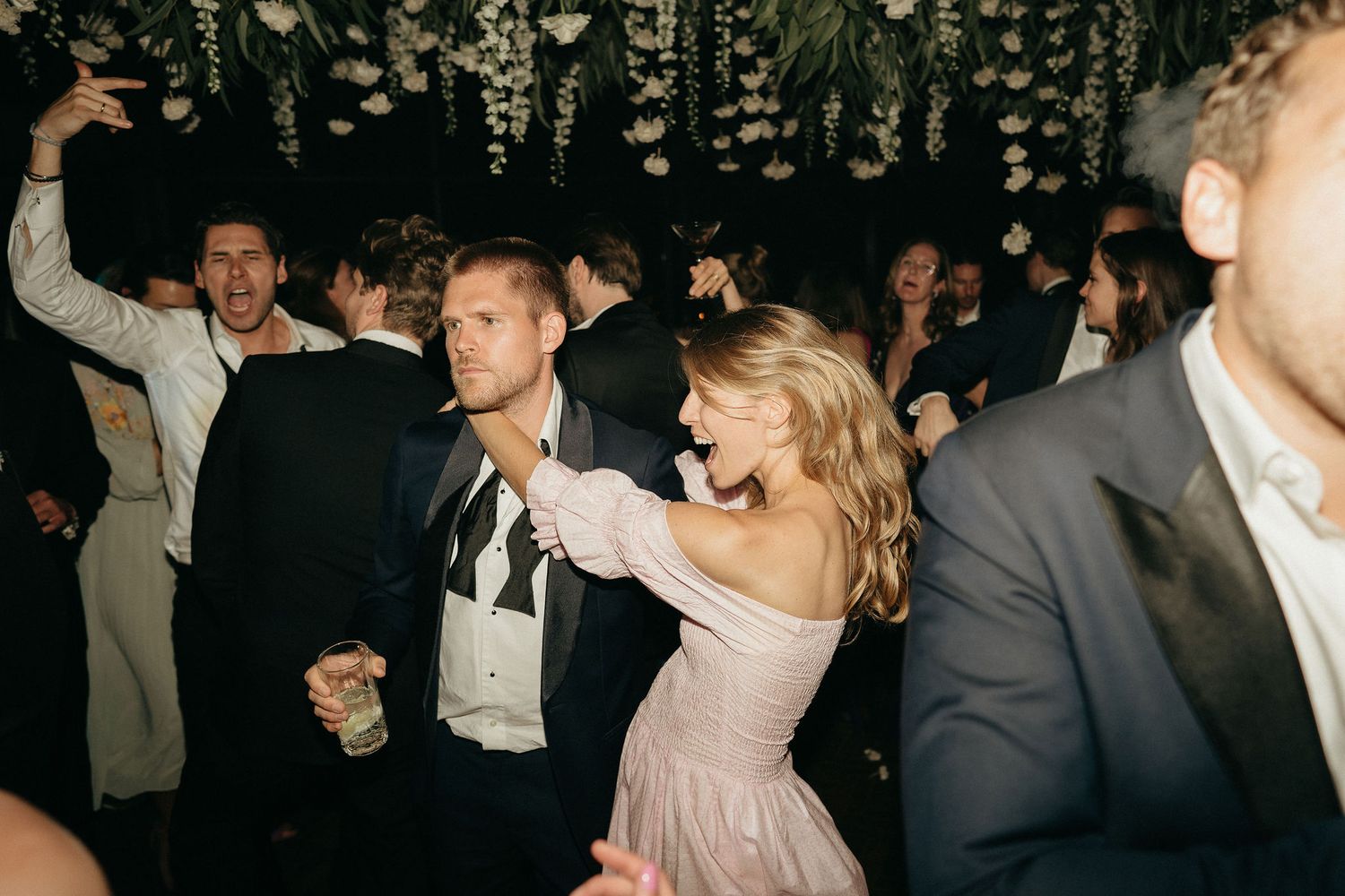 A group of people dancing and celebrating at a wedding reception in a dimly lit venue.