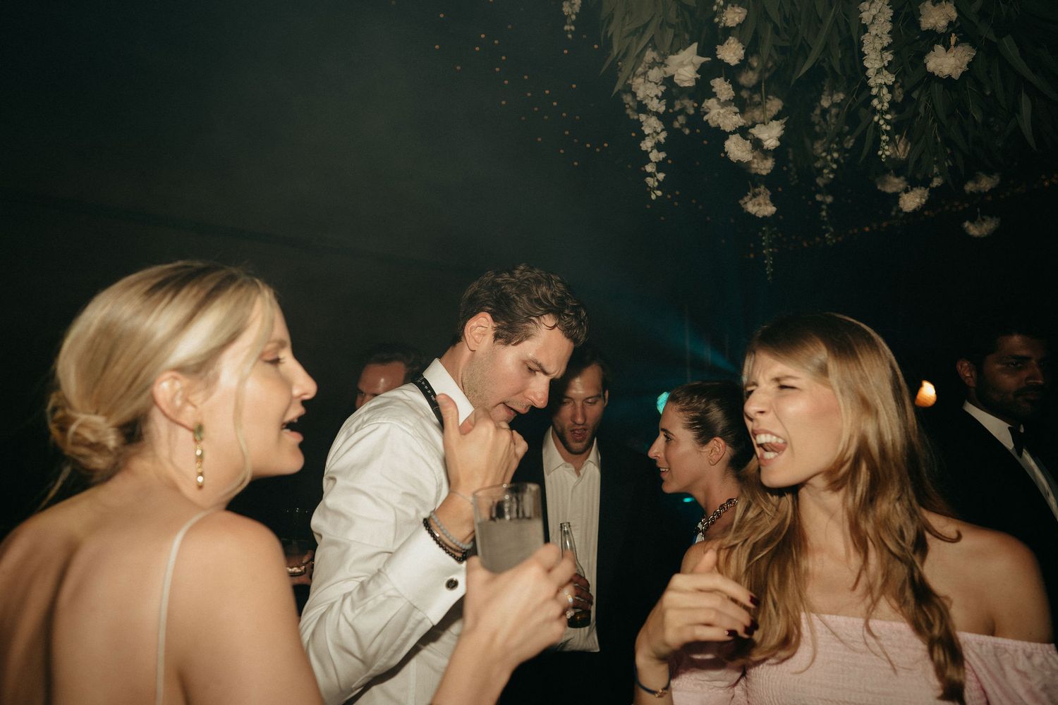 Party guests dancing and laughing together at a dimly lit celebration with decorative lights hanging overhead.