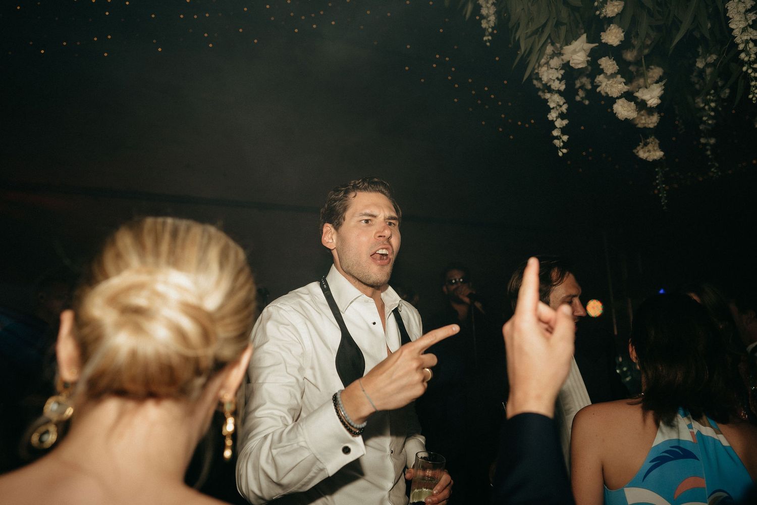 A person in a white dress shirt and tie gestures while dancing at a dimly lit party or reception.