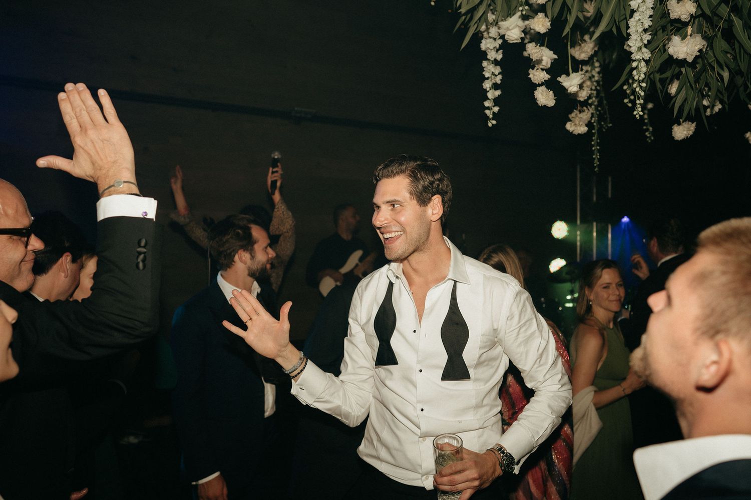People dancing and celebrating at a wedding reception with decorative lighting in the background.