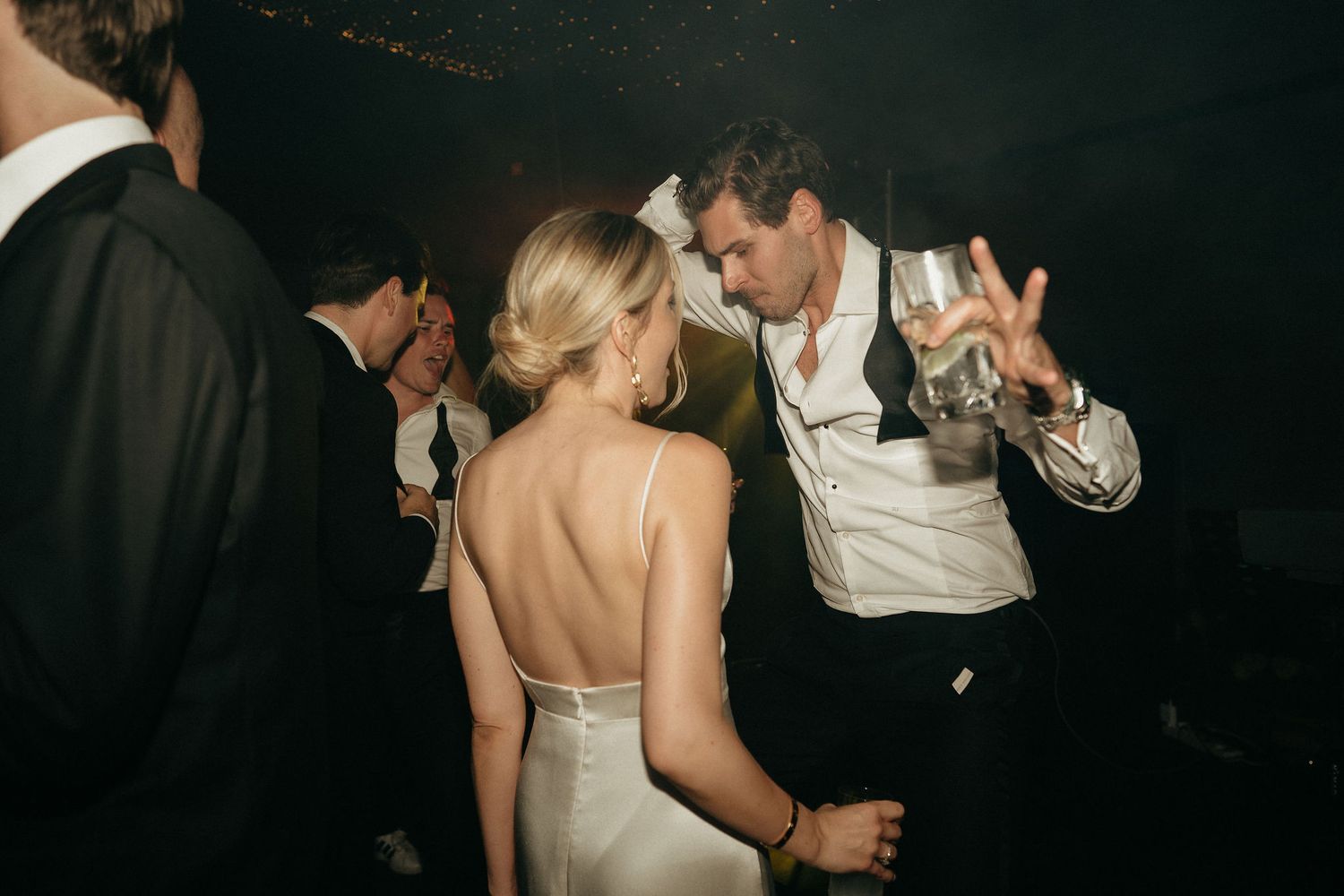Wedding guests dance and celebrate together on a dimly lit dance floor during an evening reception celebration.