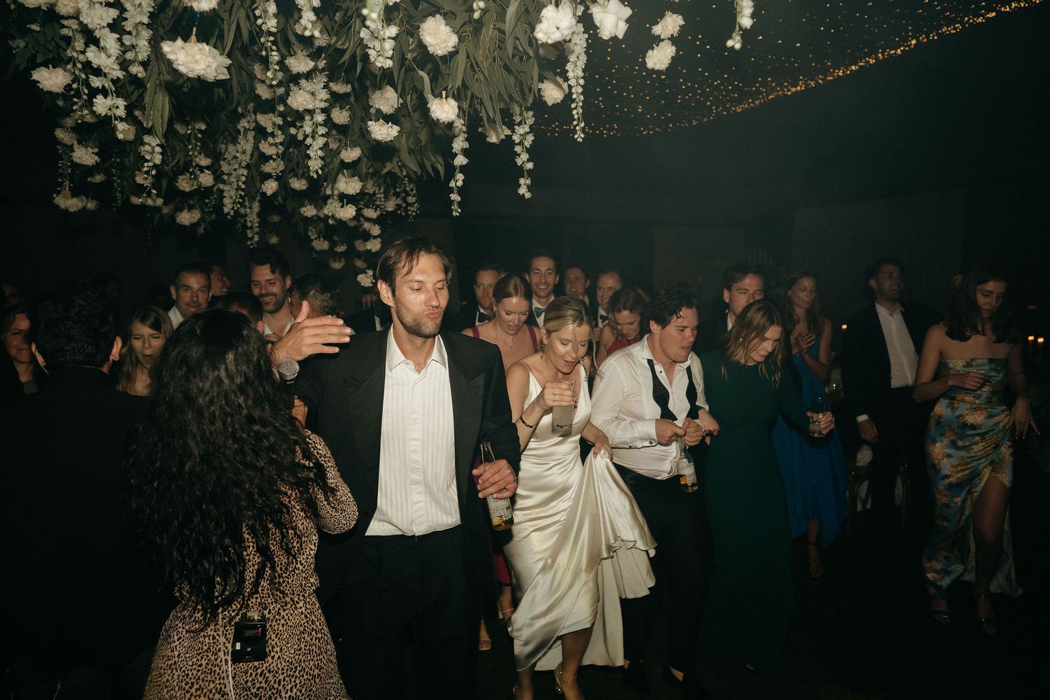 People dance together in a dimly lit reception hall under elegant chandeliers during a wedding celebration.