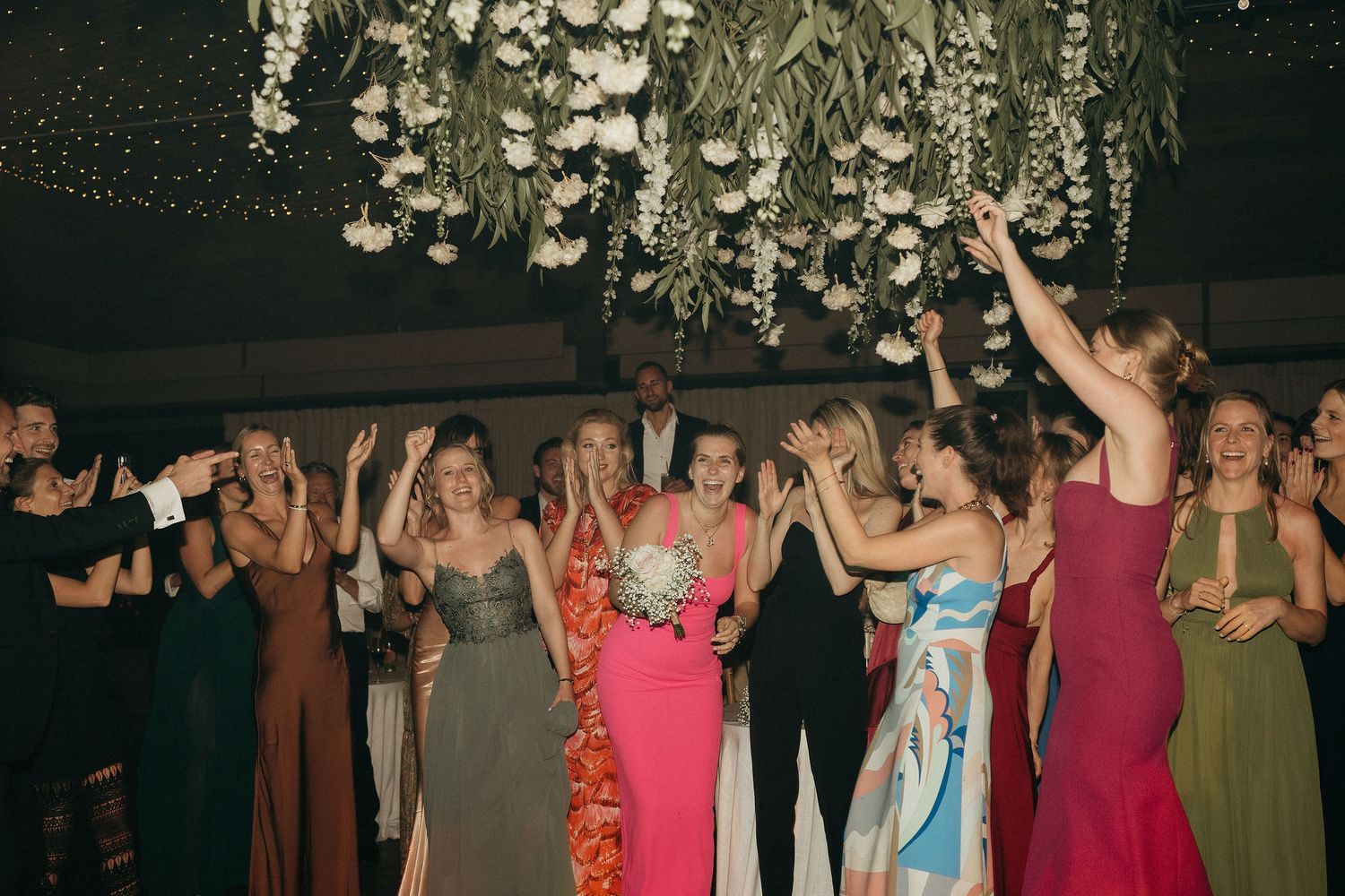 A group of formal event attendees dancing and celebrating under decorative hanging lights at a party.
