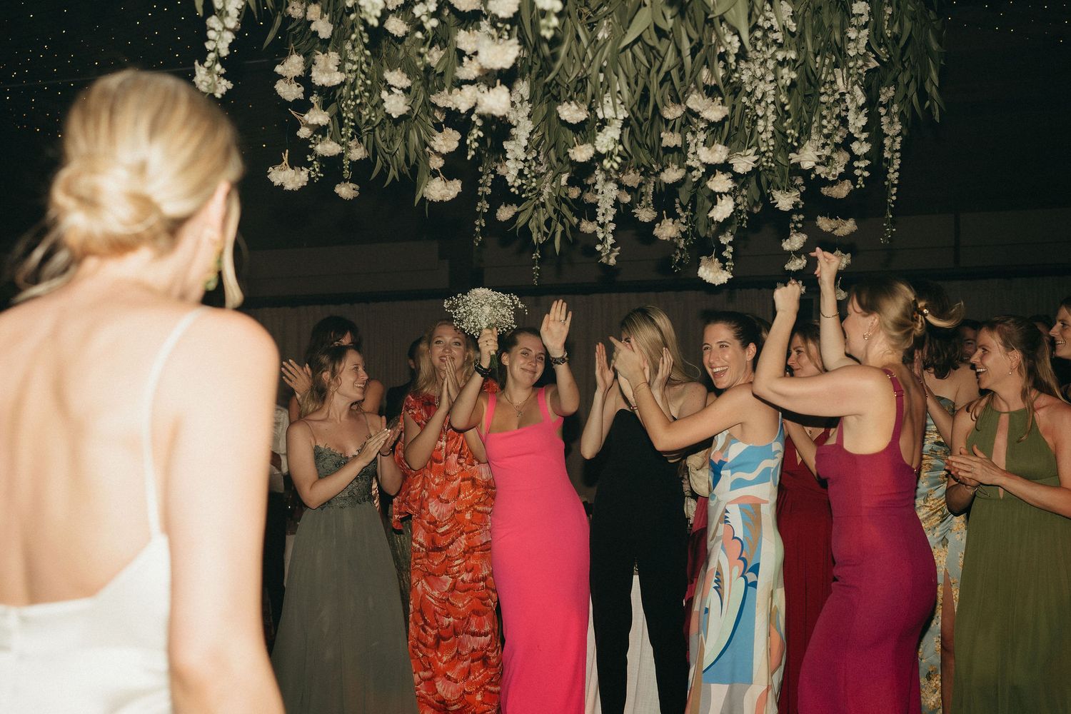 A group of people in formal attire dancing and celebrating under an ornate crystal chandelier at an evening event.