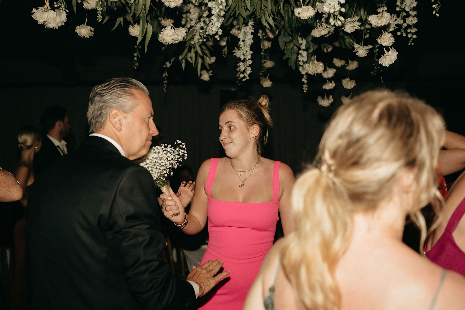 Wedding guests dance together on a dark dance floor with a pink dress visible in the background.