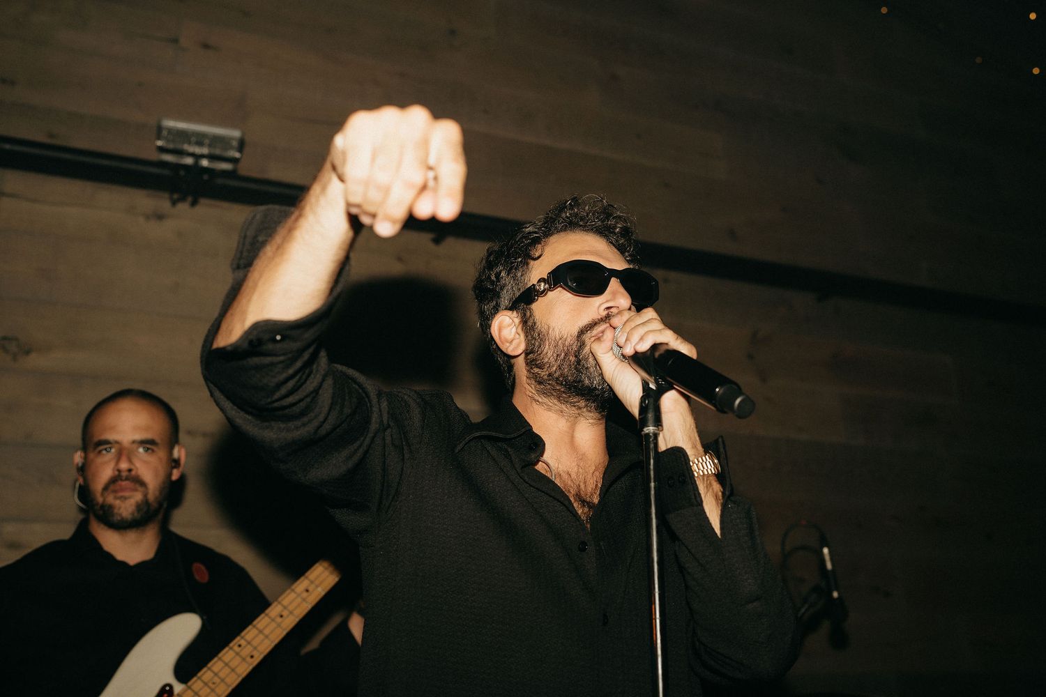 A performer in dark clothing sings passionately into a microphone on a dimly lit stage.