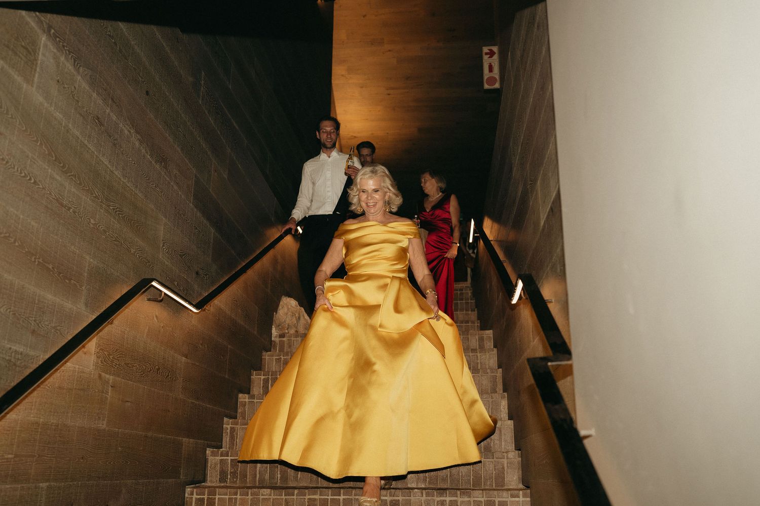 A person in a yellow formal gown descends a dimly lit staircase with dramatic lighting and stone walls.
