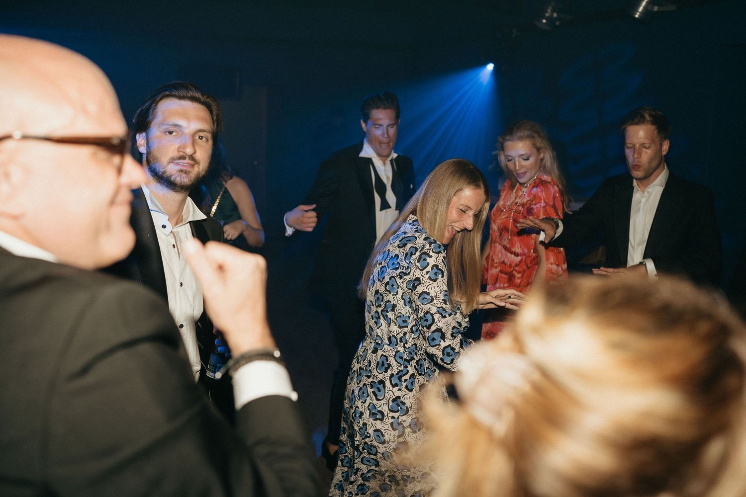 A group of people in formal attire dancing and socializing at an evening event with blue lighting in the background.