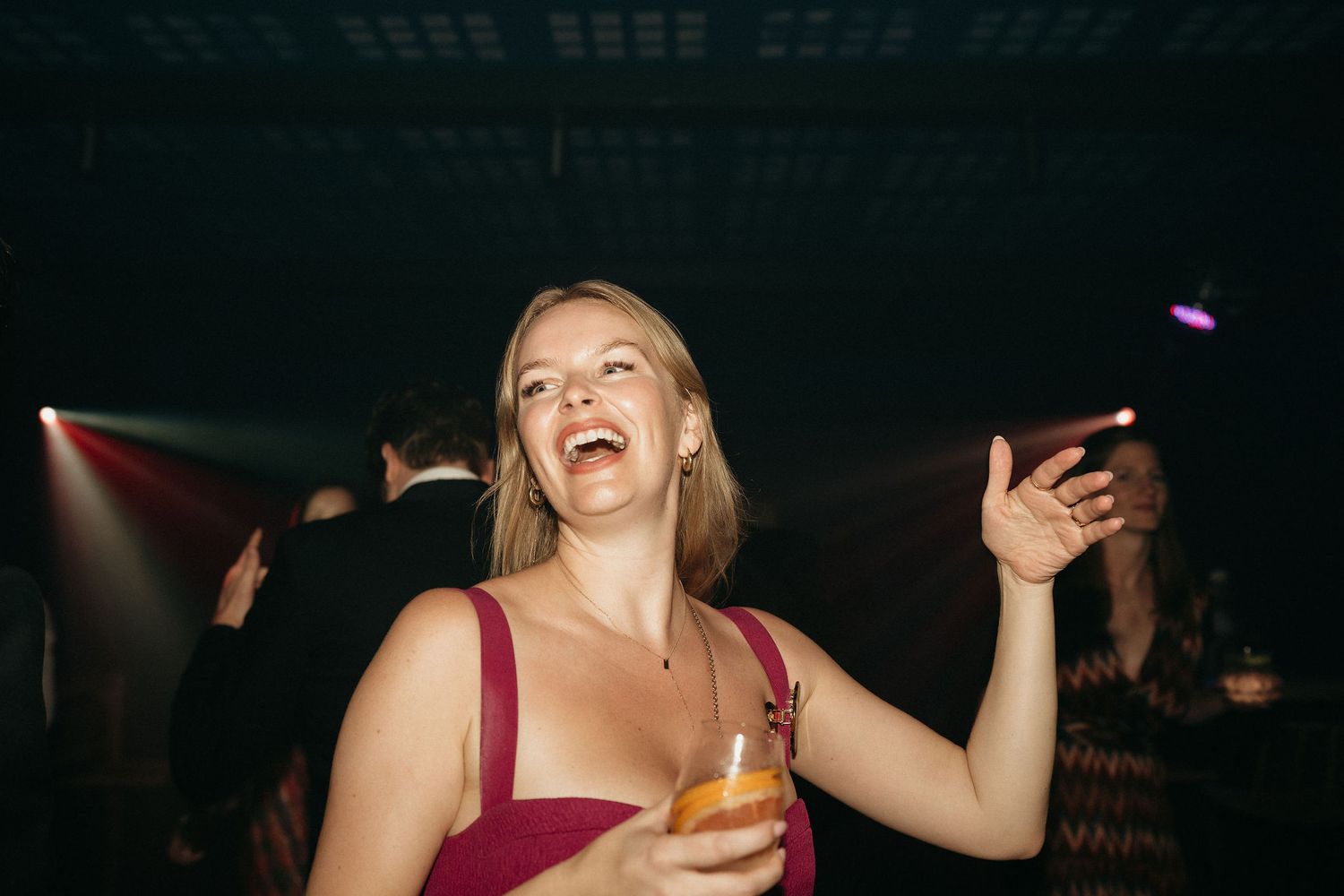 Someone in a burgundy dress dances joyfully at a nightclub while holding a drink in a dimly lit atmosphere.