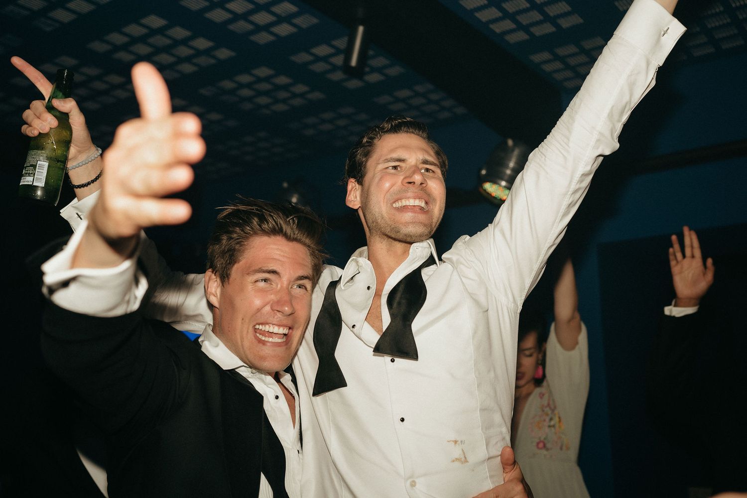 A group of people dancing and celebrating with raised arms in a dimly lit venue.