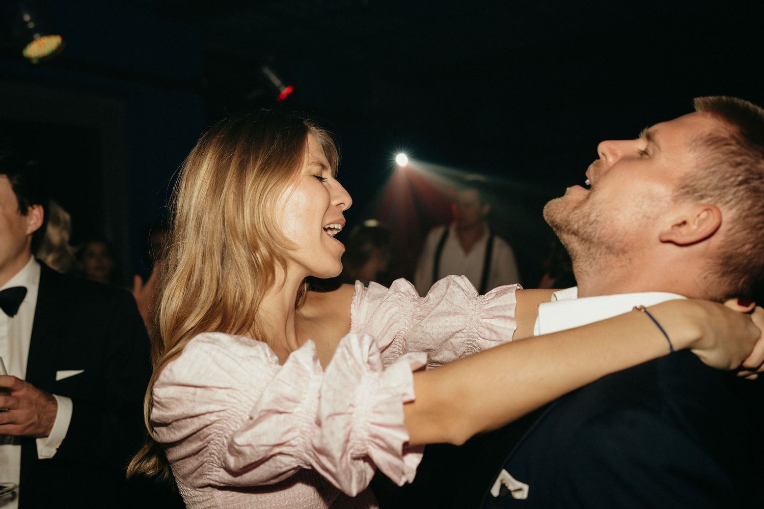 A couple dancing and laughing together at a dimly lit party, with the woman wearing a light pink ruffled top.