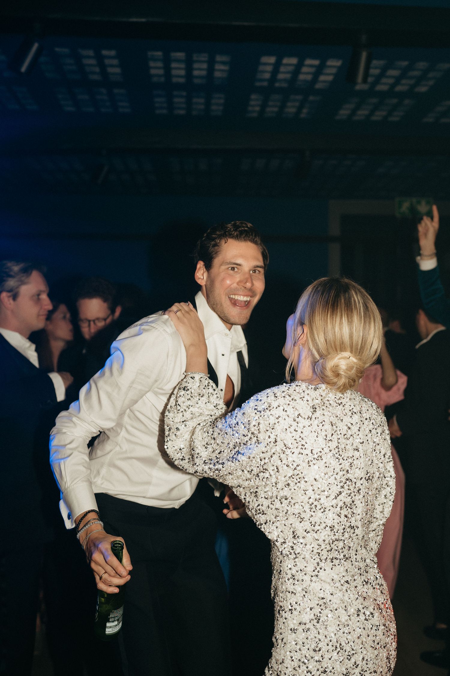 People dancing together at a dimly lit party, with someone wearing a white jacket and another in a speckled dress.