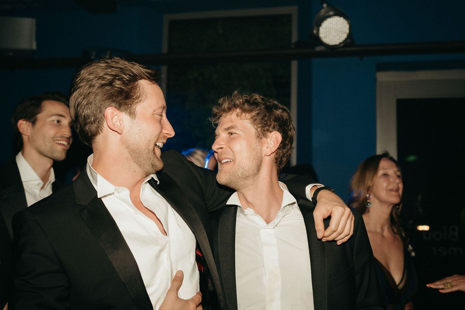 Two men in white shirts and dark jackets share a laugh together at what appears to be an evening social event.