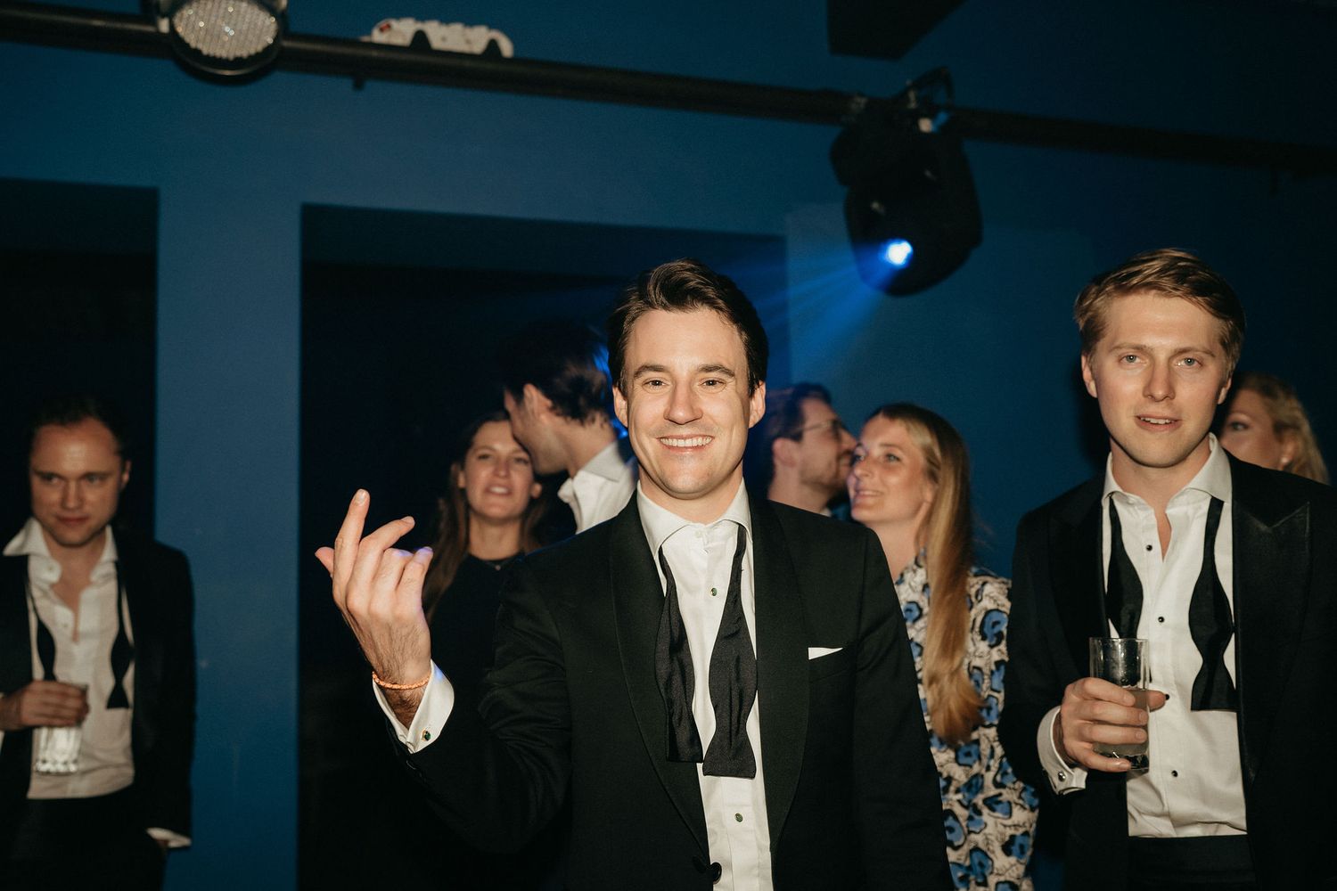 People in formal attire gathered at an event with dark blue lighting and decorative elements in the background.