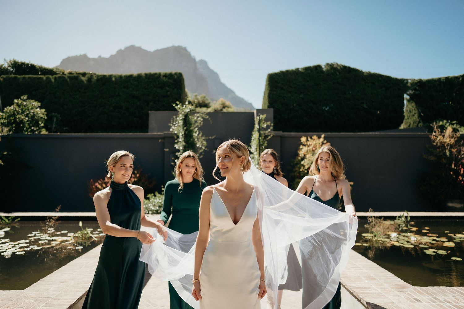 Wedding party celebrates and dances together outdoors with mountains and blue sky in the background.