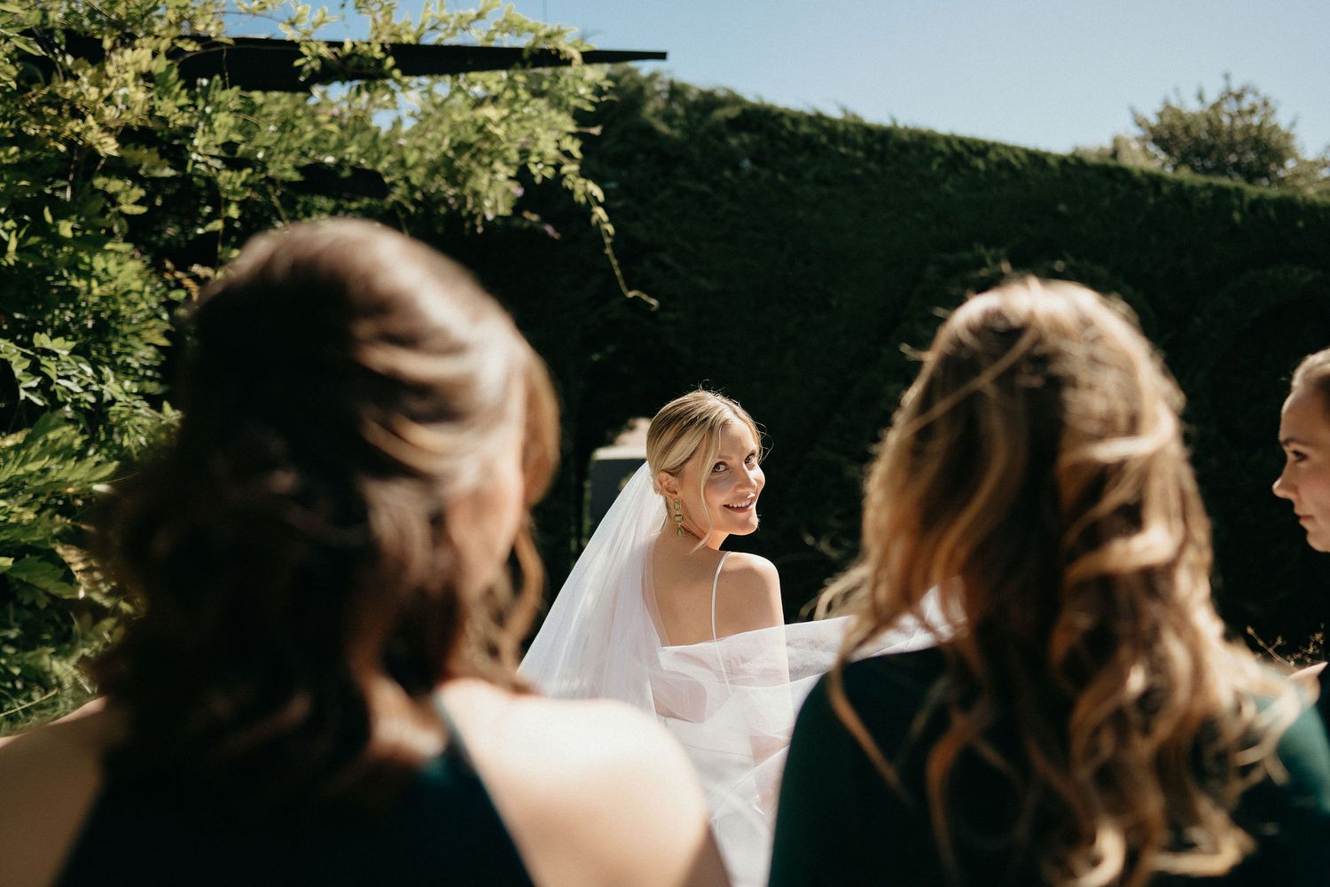 A wedding party dances and celebrates outdoors on a sunny day surrounded by greenery.