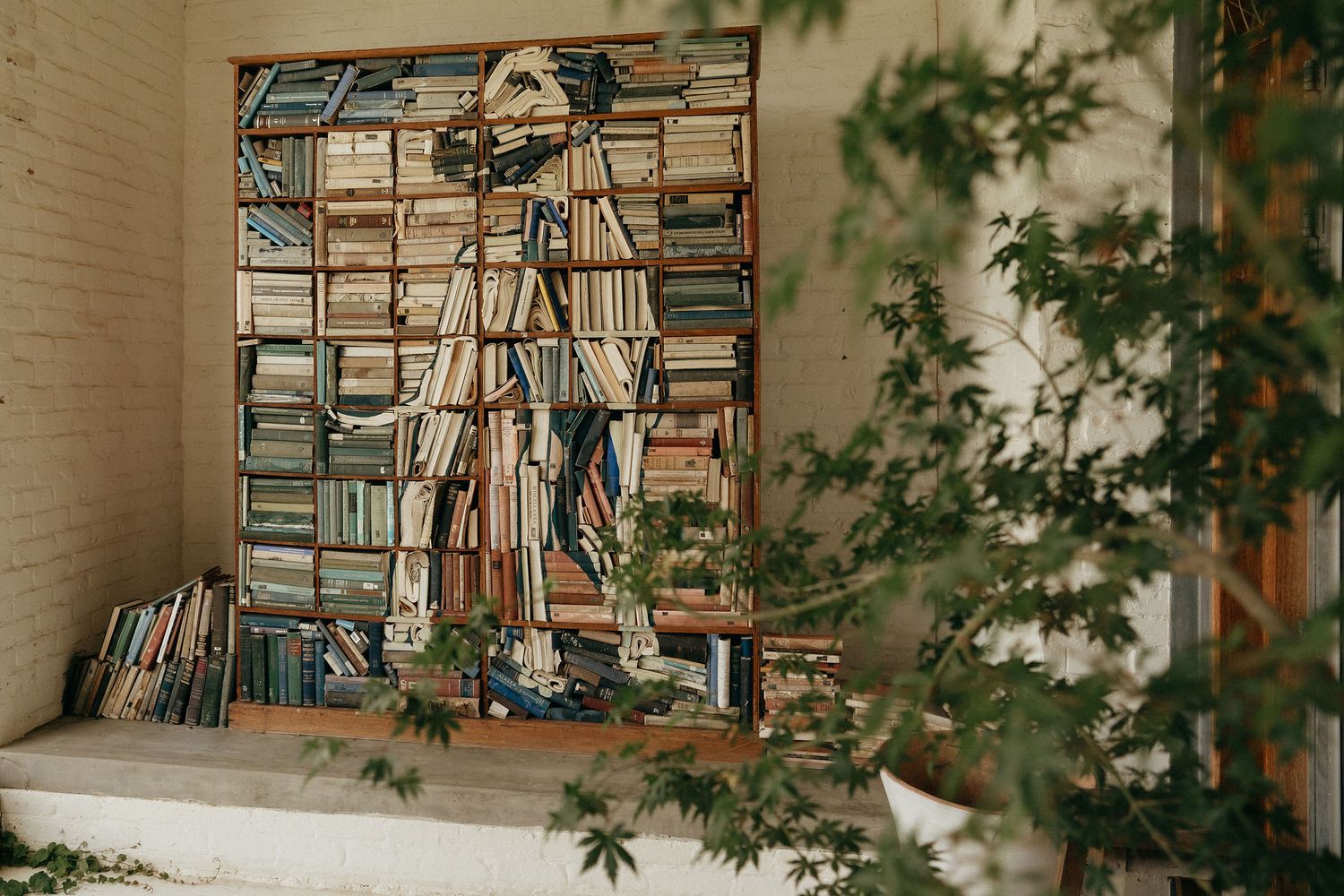 Someone stands on a ladder organizing books on tall wooden shelves filled with stacks of literature and papers.
