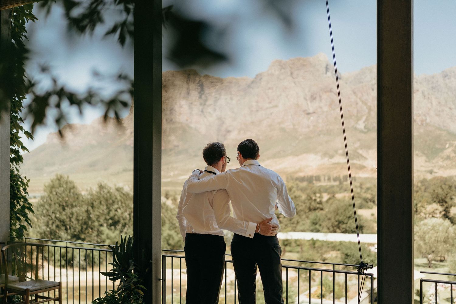 Two people stand on a balcony overlooking a desert landscape with mountains and palm trees in the distance.