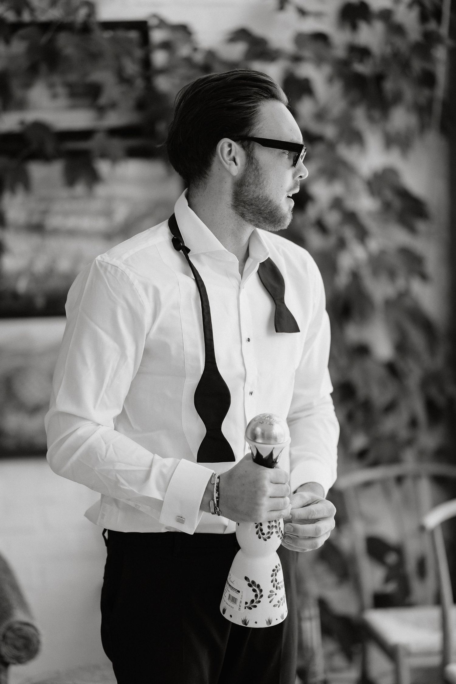 A person in a white dress shirt and black tie holds a wine glass in an outdoor setting.