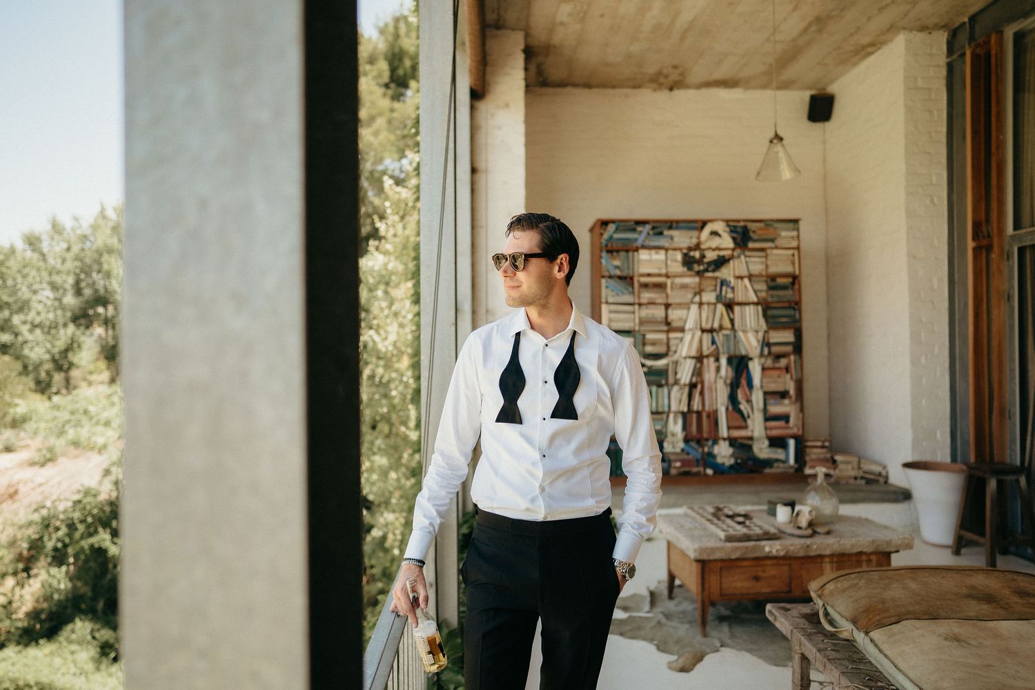 A stylish groom in a white shirt and black suspenders stands in a rustic room with natural light streaming through windows.