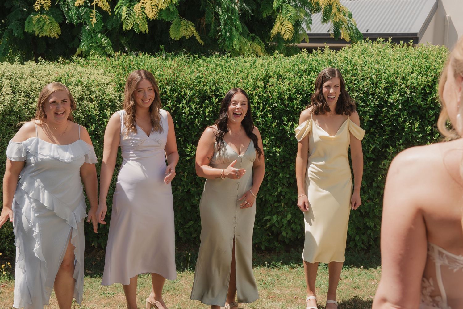 A row of people wearing neutral-toned dresses stand together outdoors against a green garden background.