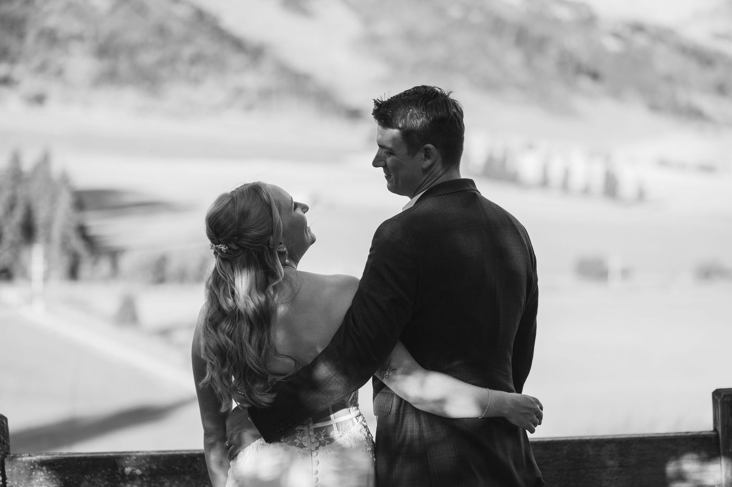 A couple in winter attire embraces while looking at a snowy mountain landscape in black and white.