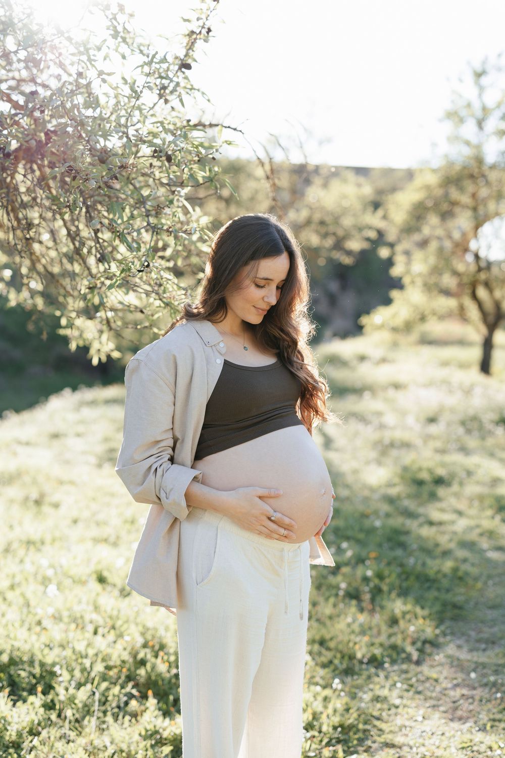 Pregnant woman in beige outfit cradling her baby bump outdoors in a sunny green garden.