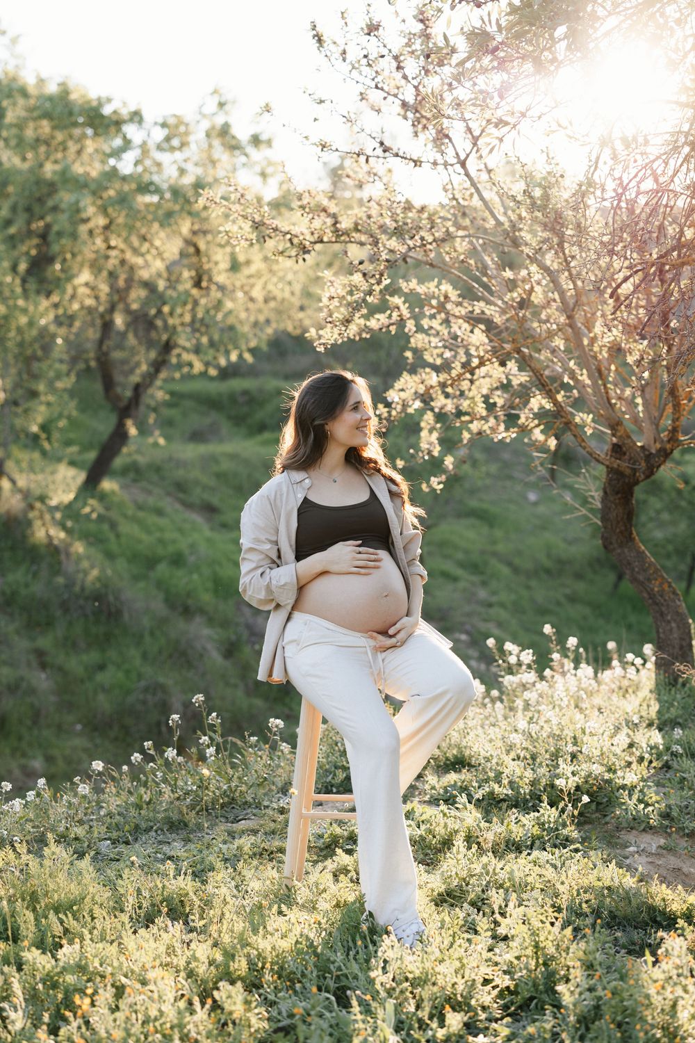 Pregnant woman sitting on stool outdoors near blooming trees in golden sunlight, cradling her baby bump in white pants.