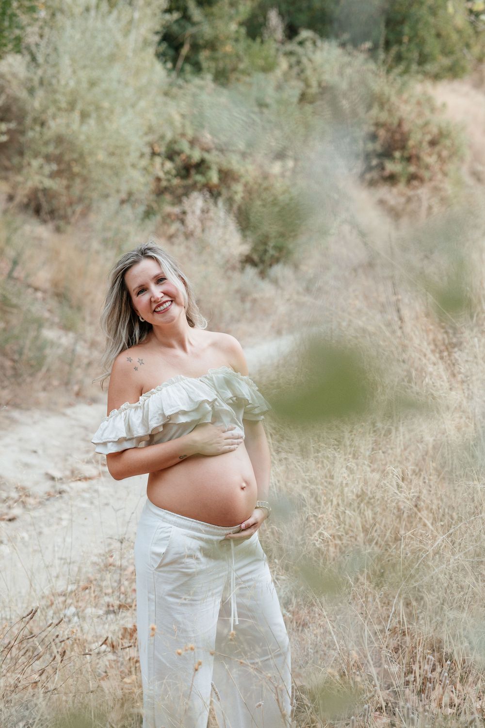 Candid maternity photos of expecting mother in white dress standing in sunlit field.