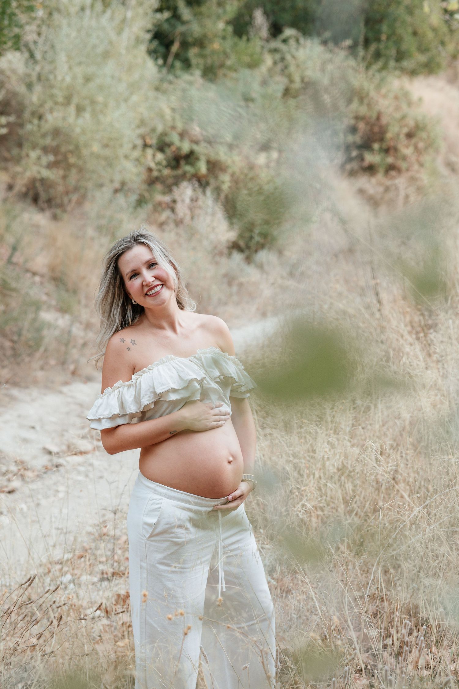 Candid maternity photos of expecting mother in white dress standing in sunlit field.