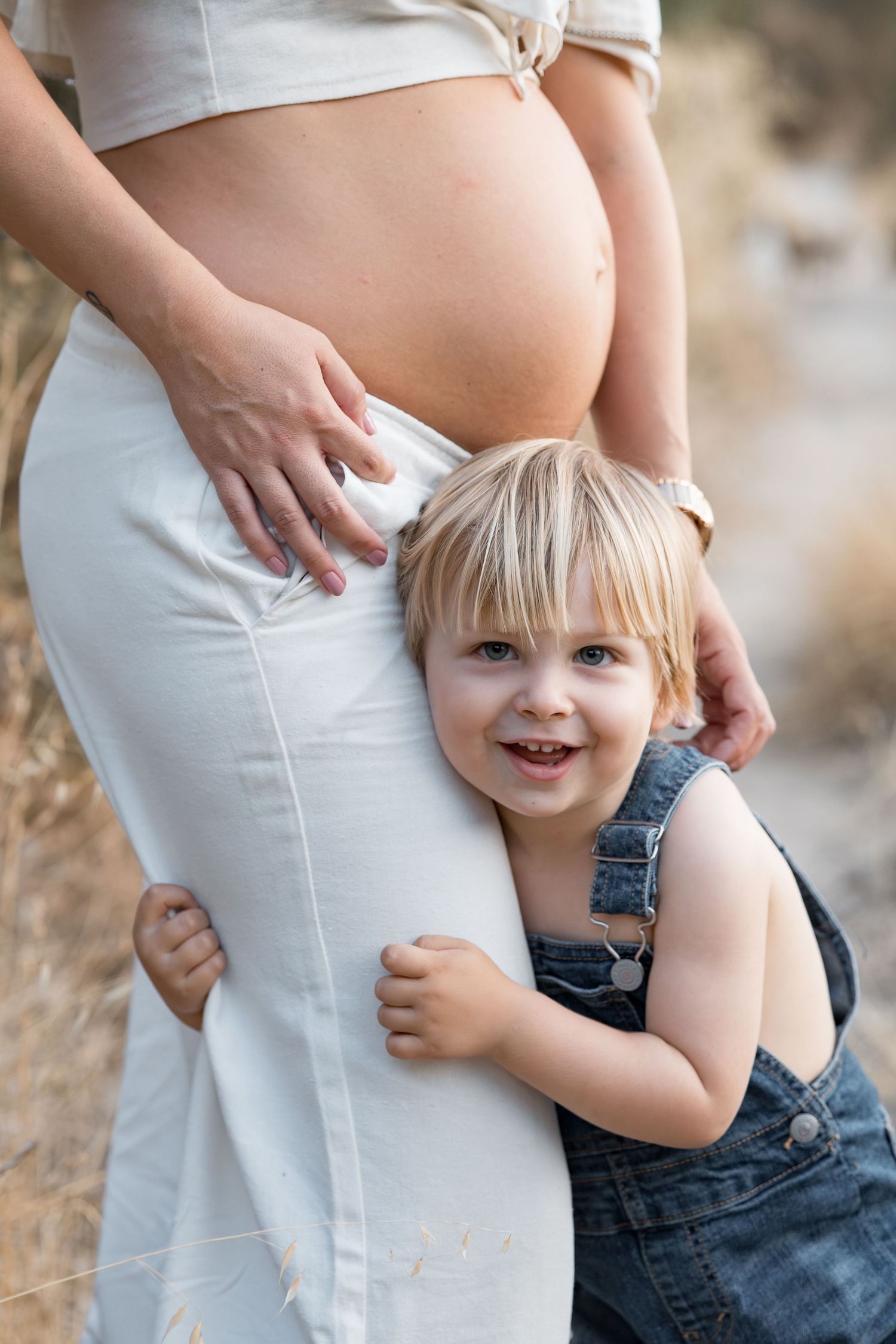 Maternity photo showing pregnant woman in white dress with toddler hugging her belly.