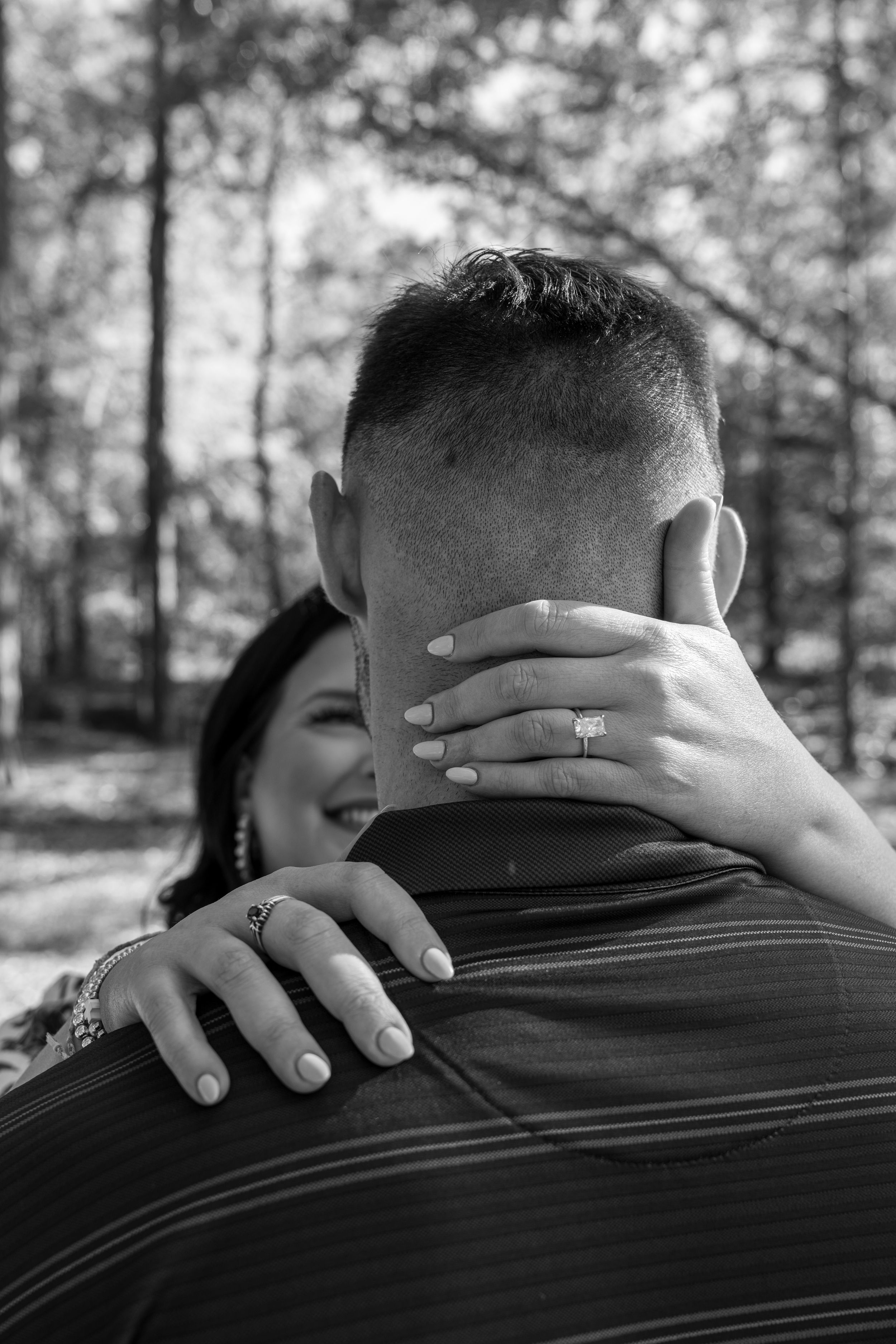 Black and white photo of a couple embracing outdoors, woman smiling with engagement ring visible on her hand.