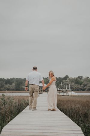Couple holding hands on a wooden dock overlooking a marsh and river on a cloudy day.