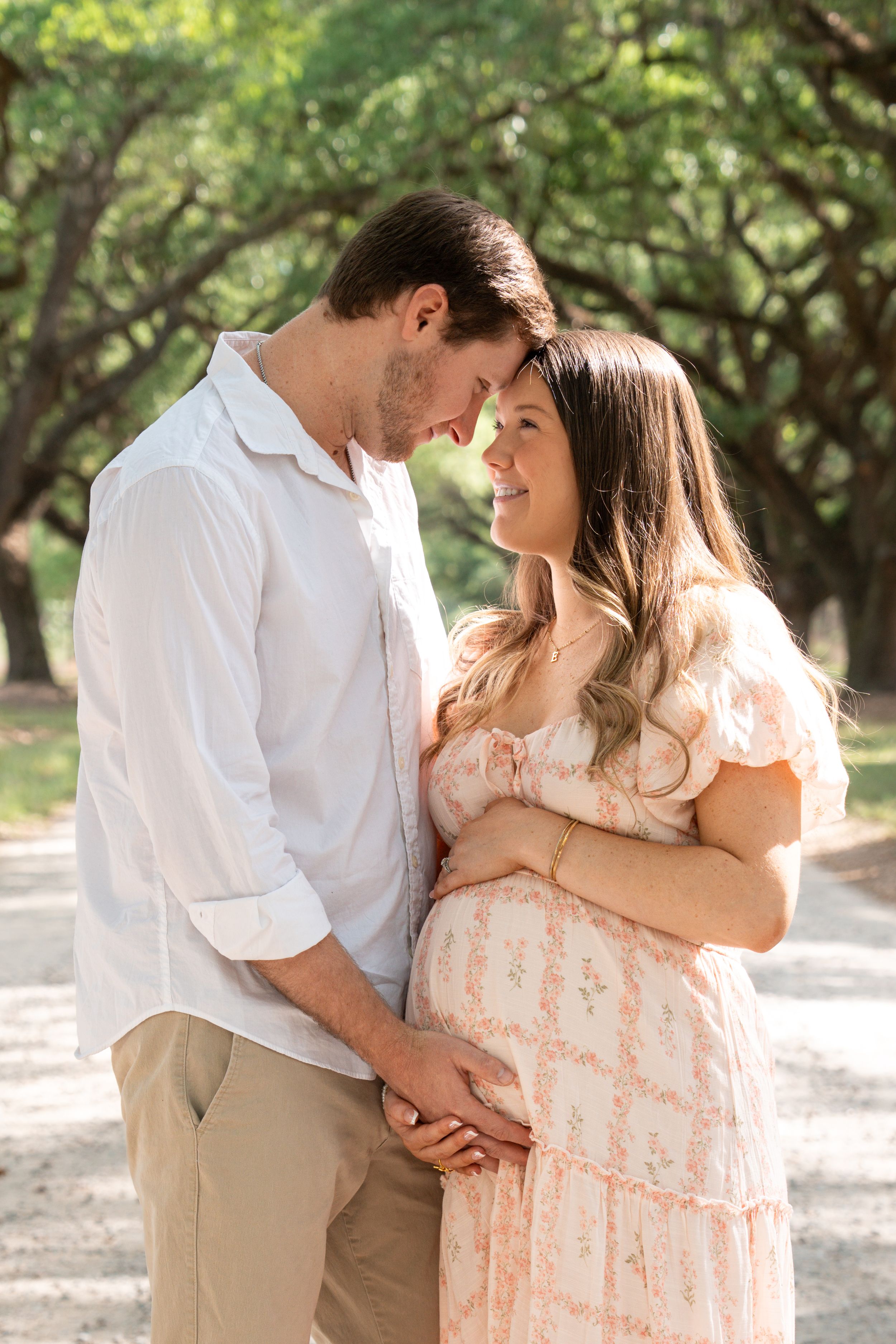 A couple shares a tender moment outdoors, the woman in a floral dress cradling her baby bump while her partner leans in close.